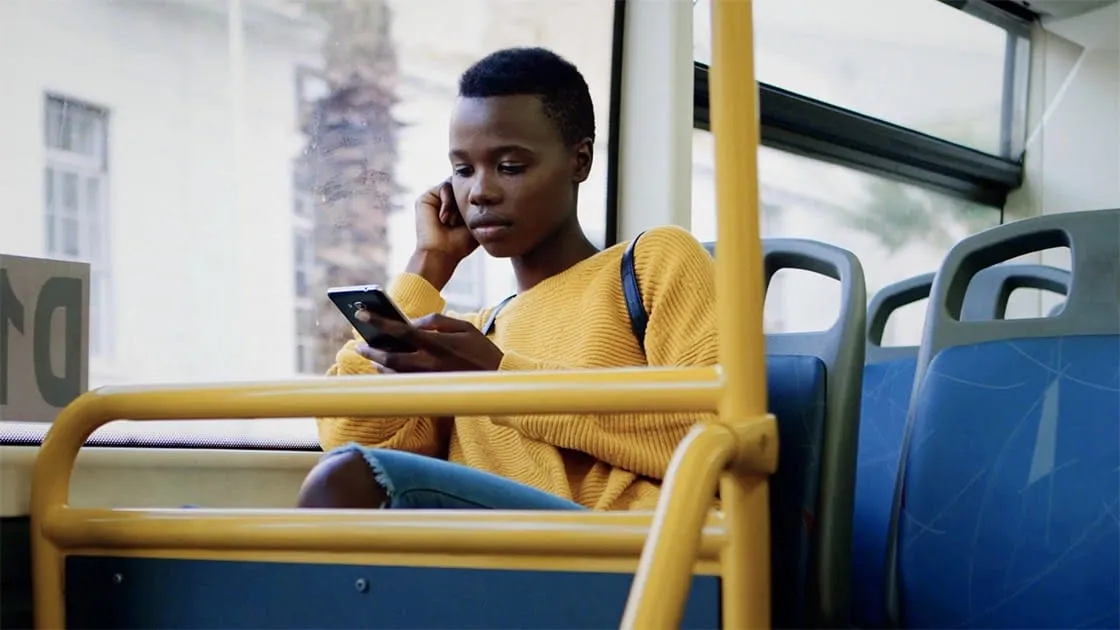 A young woman in a yellow jumper sits on a bus, looking at her phone.