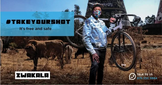 A man in a face mask stands with his bicycle beside grazing cattle, under the words “#TakeYourShot: it’s free and safe”
