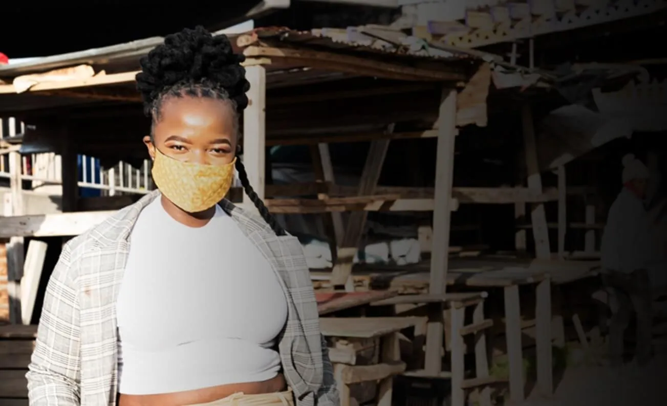 A young woman in a yellow patterned face mask stands in front of wooden market stalls.